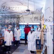 Researchers pose in their lab behind glass with the words "COVID Warriors" written on it.